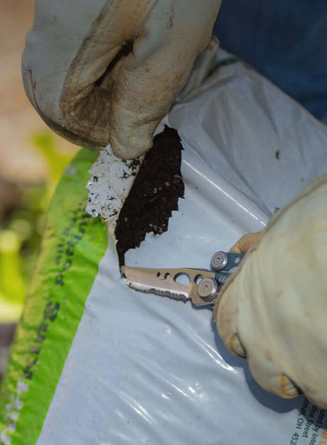 Person using a Denim Skeletool® to open a bag of soil, showcasing the utility of this versatile tool in gardening tasks.