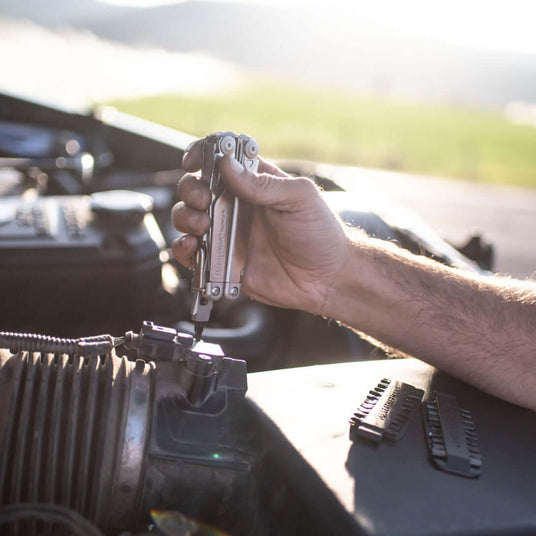 A hand using a stainless steel Leatherman Surge® multitool on a car engine, showcasing its practical use in repairs.