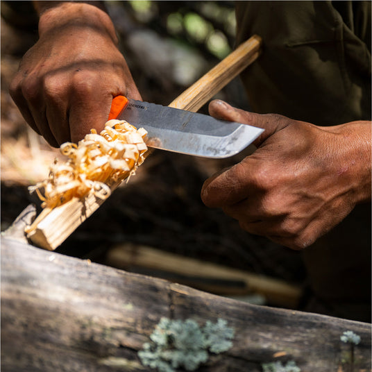 Close-up of hands using an Orange Pop Leatherman Pioneer™ knife to carve wood shavings outdoors.