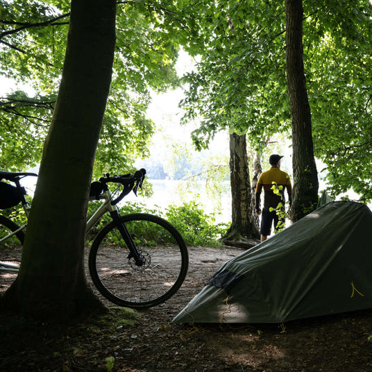 A cyclist stands near a tent by the river, surrounded by lush trees in a tranquil camping setting.