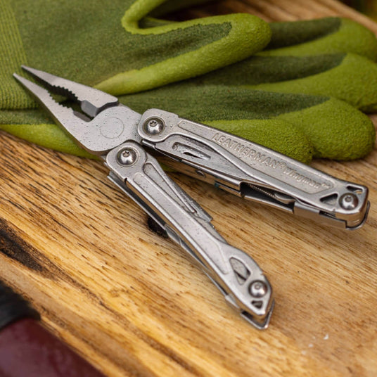 Stainless Steel Leatherman Wingman® multi-tool on a wooden surface beside green gloves.