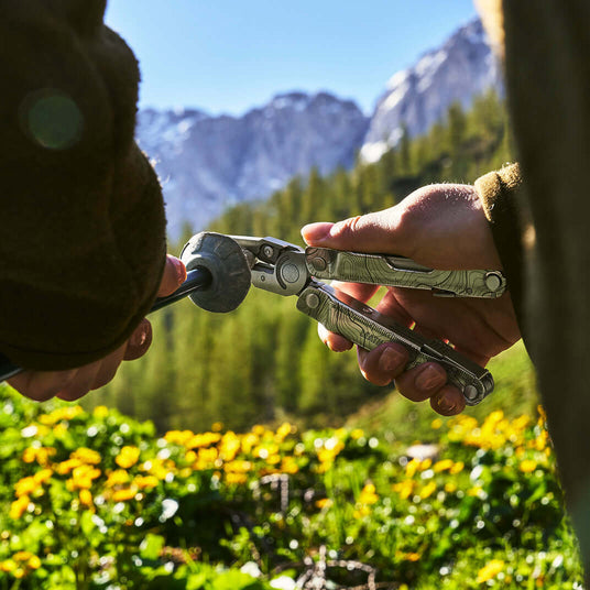 Hands using a customized Stainless Steel Leatherman Rebar® multitool outdoors with mountains in the background.