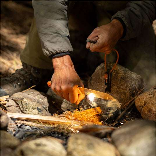 Translation missing: en.Person using a Leatherman Pioneer knife and fire starter to ignite feather sticks in a campfire setup, shown in an outdoor setting with sparks and smoke rising from the tinder