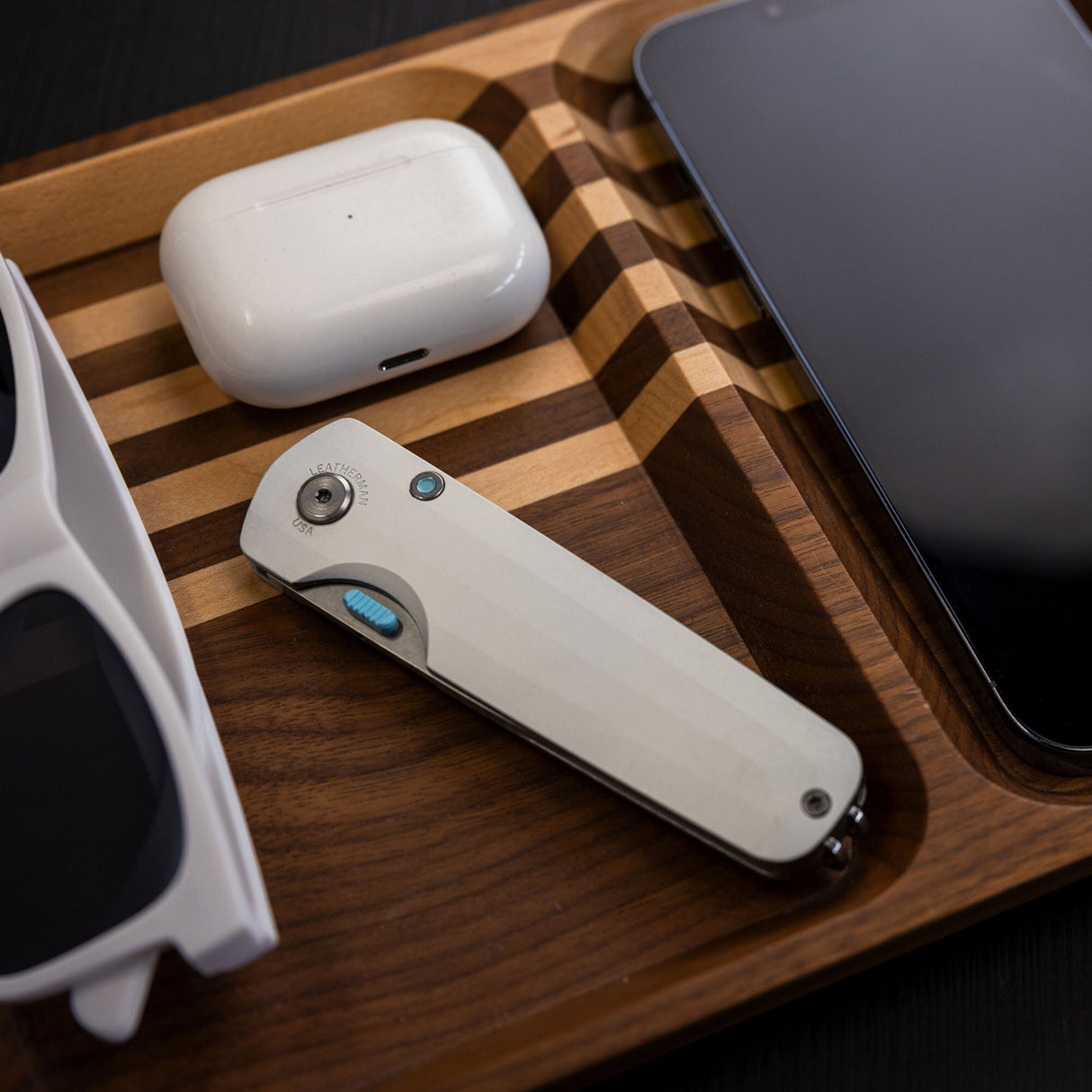 Translation missing: en.Leatherman Blazer Alpine folding knife in closed position placed in a wooden valet tray alongside wireless earbuds, a smartphone, and sunglasses, highlighting everyday carry essentials