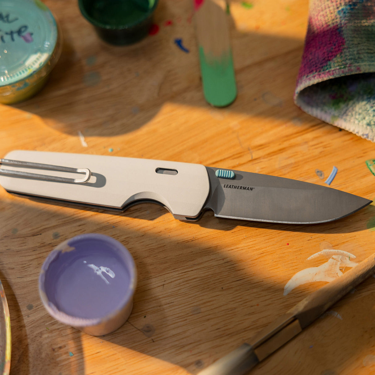 Translation missing: en.Leatherman Blazer Alpine folding knife shown open on a wooden table surrounded by paint supplies and a brush, highlighting its role as a creative or everyday utility tool