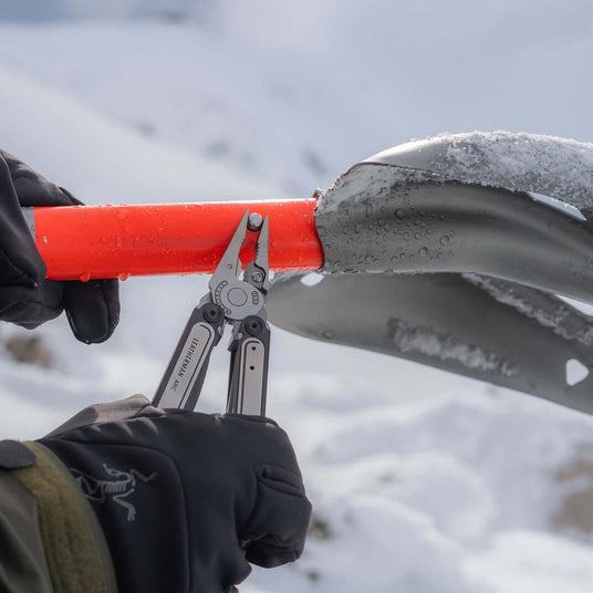 Person using a Black DLC & Stainless Steel Leatherman ARC® multitool on a snowy shovel.
