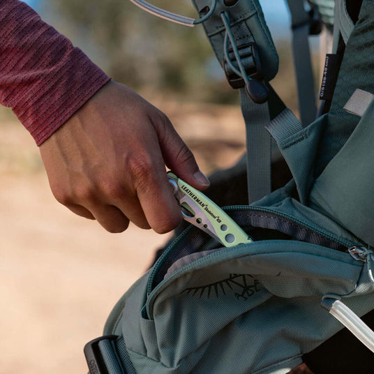 Hand reaching into backpack pocket to retrieve a Verdant Leatherman Skeletool® KB for outdoor activities.