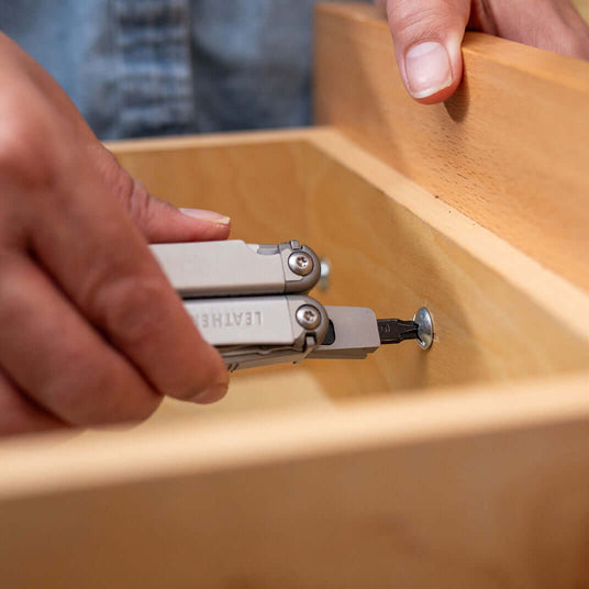 Hand using a Stainless Steel Leatherman Wave® Plus multitool on a wooden drawer, showcasing its practical features.