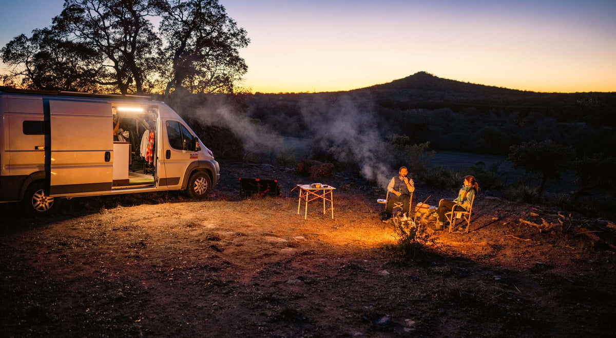 Campers enjoying a fire beside a van at sunset in a scenic outdoor location.