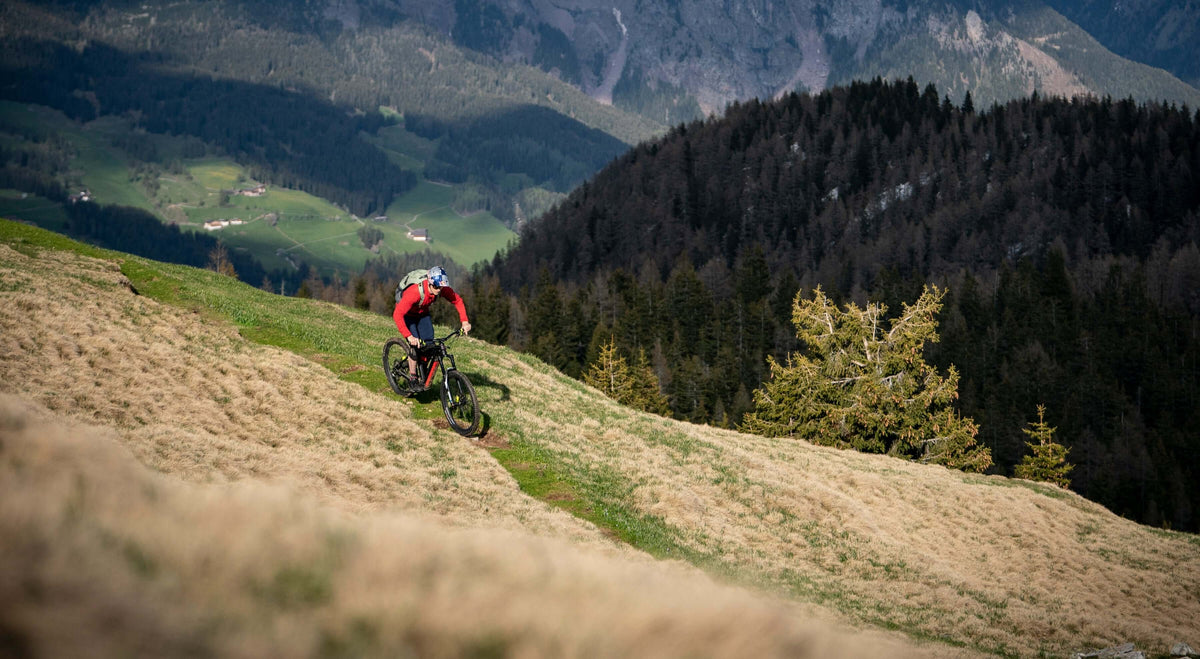 Mountain biker riding on a trail with scenic mountain views and lush greenery in the background.