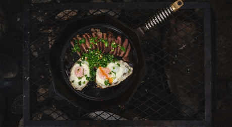 A cast iron skillet on a grill, featuring sunny-side up eggs, sliced meat, and fresh herbs.