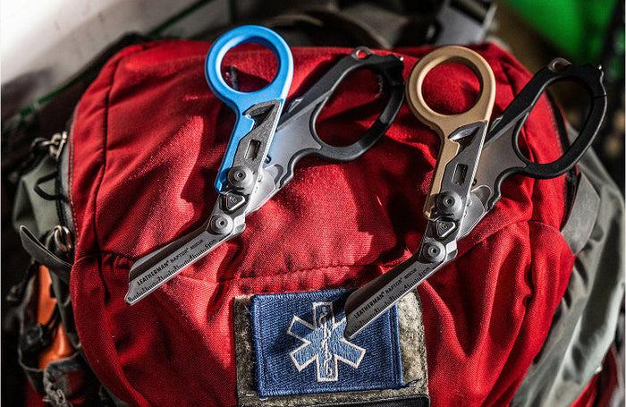 Translation missing: en.Two Leatherman Raptor Rescue shears—one with a blue/blue handle and the other with a tan/black handle—rest on a red first aid backpack adorned with a blue medical star patch, emphasizing their use in emergency medical or rescue situations