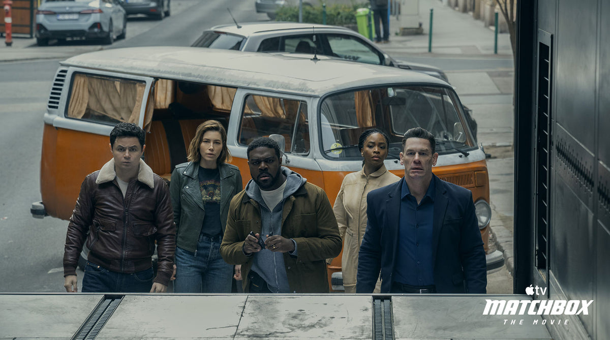 Translation missing: en.Five actors walk forward in front of a vintage orange and white van on a city street, with cars and a sidewalk visible in the background. Matchbox The Movie Apple TV logo appears in the bottom right corner