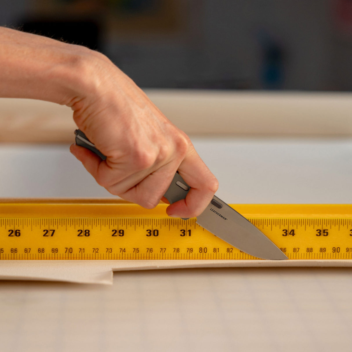 Translation missing: en.Person using a Leatherman Blazer Stainless Steel folding knife to cut fabric along a yellow measuring ruler on a gridded work surface, shown in a close-up of the hand and cutting action