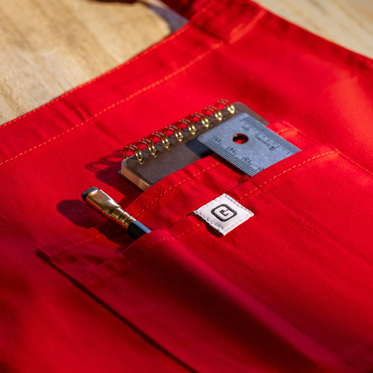 Translation missing: en.Red apron front pocket holding a pencil, small notebook, and metal ruler
