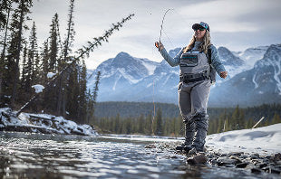 Translation missing: en.A woman stands in shallow water, casting a fly rod, surrounded by nature and reflecting a peaceful fishing scene.
