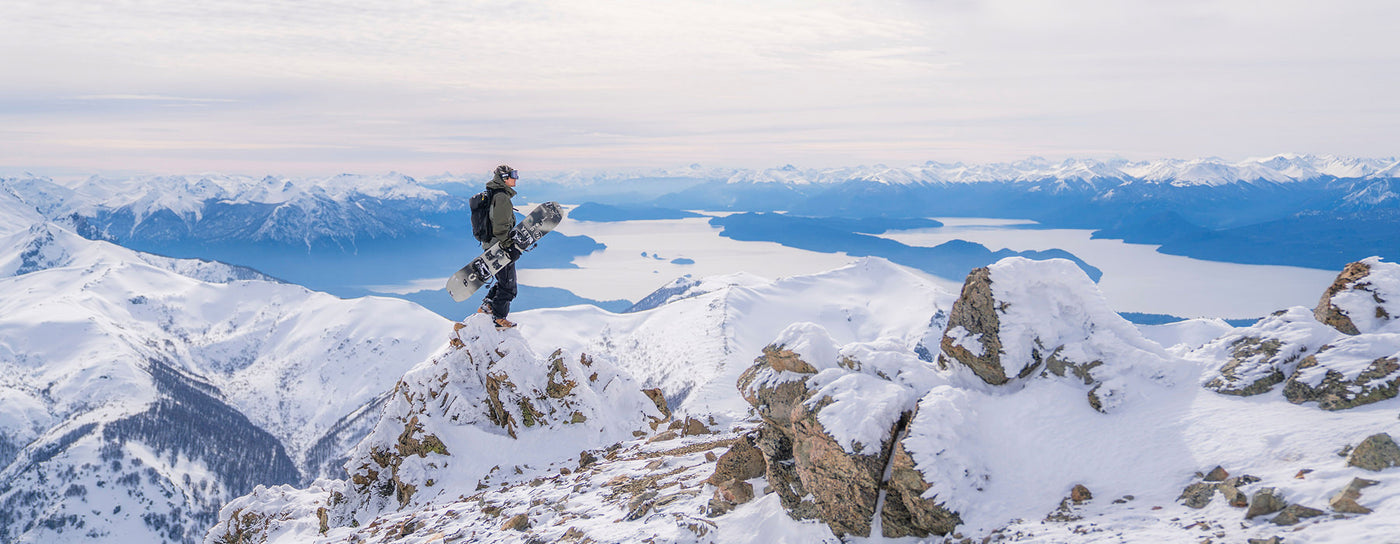Translation missing: en.A man stands triumphantly on a mountain peak, holding a snowboard against a backdrop of snow-covered slopes