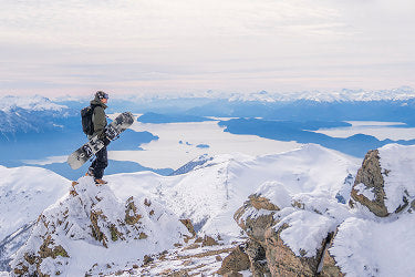 Translation missing: en.A man stands triumphantly on a mountain peak, holding a snowboard against a backdrop of snow-covered slopes