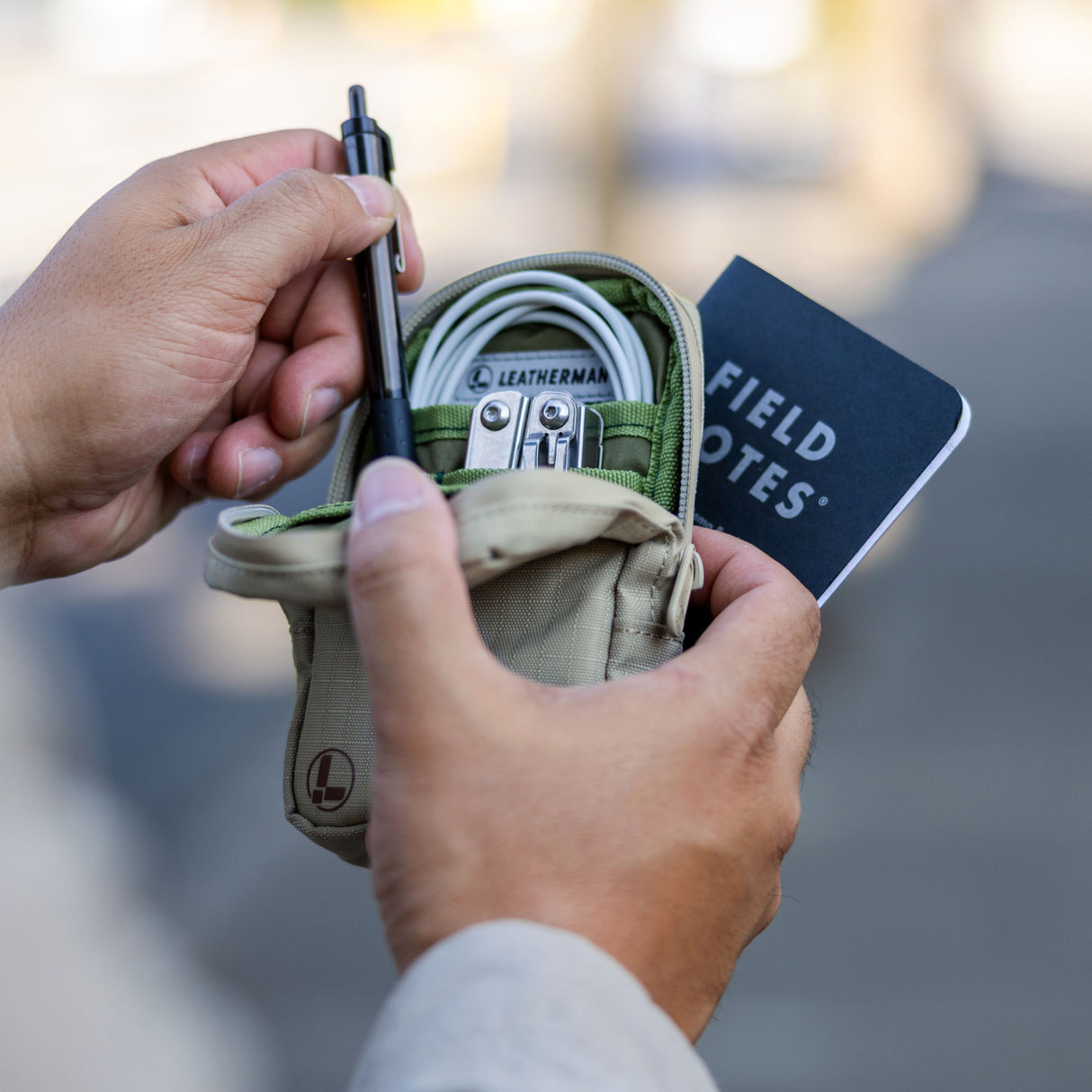 Translation missing: en.A person holds open a Leatherman Tool Pouch in Mossy Fog color, revealing neatly organized items including a Leatherman multi-tool, charging cable, pen, and Field Notes notebook—illustrating the pouch's utility for everyday carry and organization