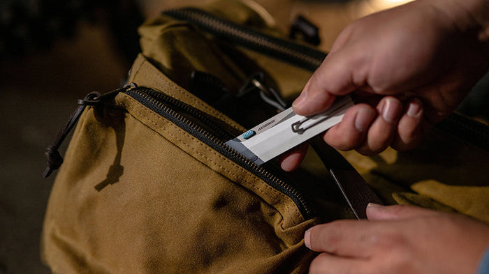 Translation missing: en.A person places or removes a Leatherman folding knife with a silver pocket clip and blue thumb stud from the front zippered pocket of a tan canvas backpack. The scene is lit warmly, emphasizing the rugged and practical nature of the gear