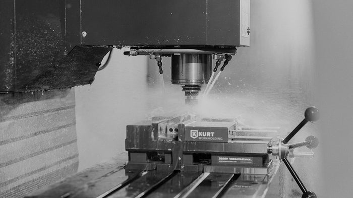 Translation missing: en.A black and white image of a CNC milling machine in operation, with coolant spraying as the machine drills into a workpiece secured in a KURT brand vise. The machining process is taking place on a metal table with linear guide rails
