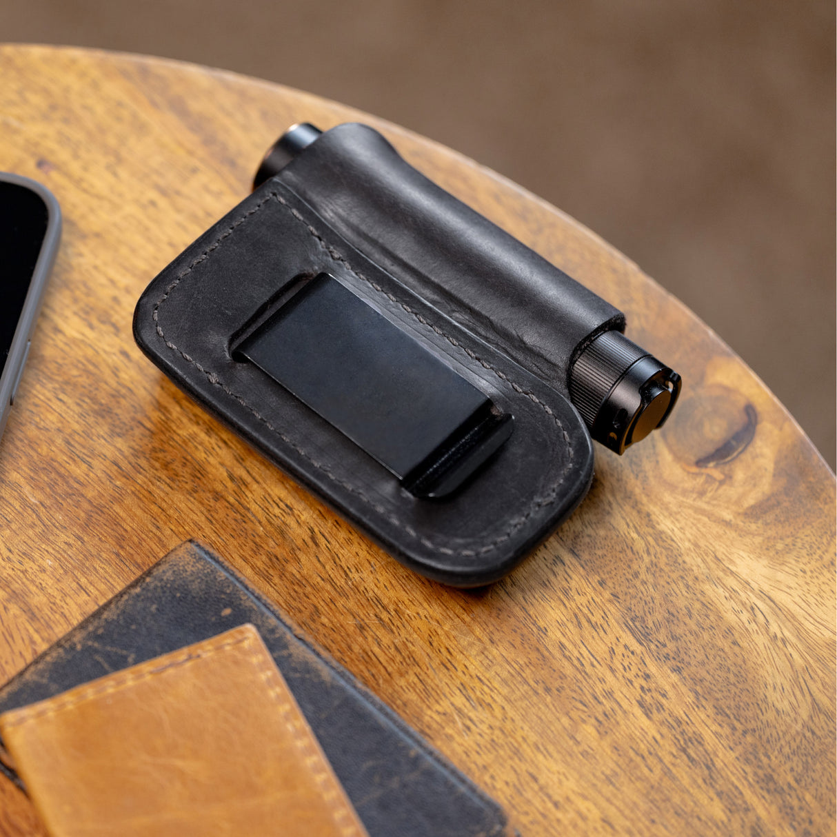 Translation missing: en.Black leather sheath with belt clip holding a small cylindrical flashlight on a wooden surface