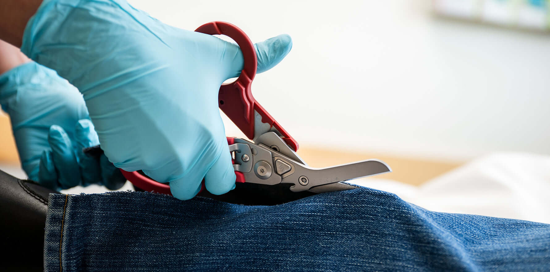 A nurse wearing blue gloves is cutting a pair of jeans with Leatherman Raptor Rescue shears, focused on the task at hand.