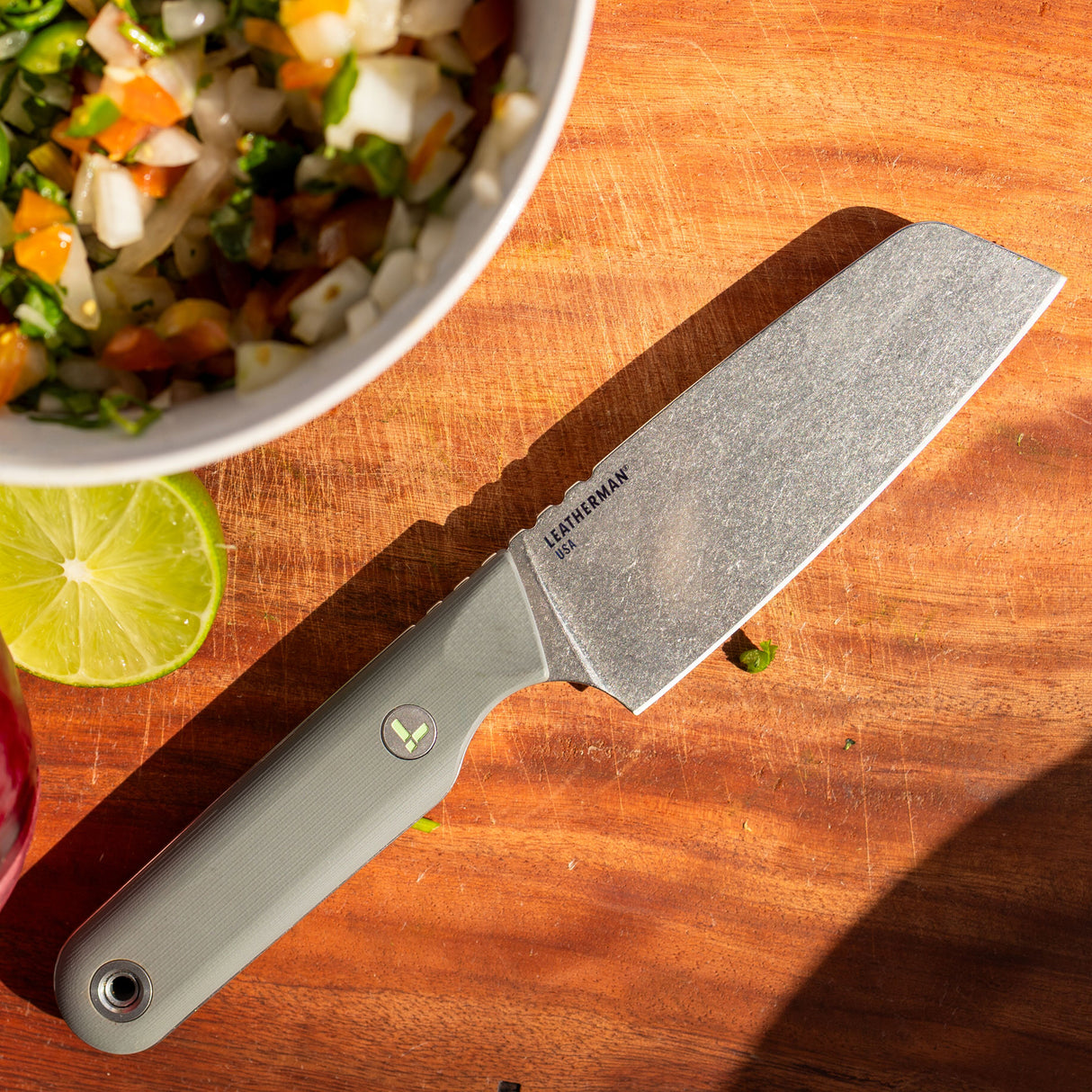 Translation missing: en.A Leatherman Rustle Vapor Grey knife resting on a wooden cutting board beside a halved lime and a bowl of freshly chopped vegetables, evoking a sense of outdoor cooking or meal prep