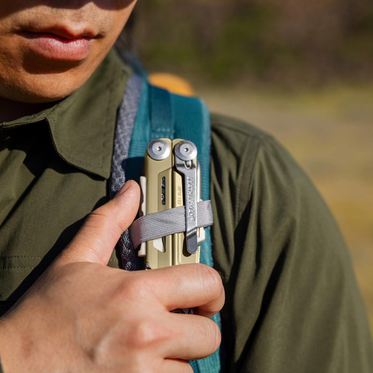 Translation missing: en.Person securing a Signal Sandstorm multitool to a backpack shoulder strap with a gray loop outdoors