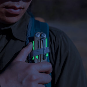Translation missing: en.Person securing a Signal Sandstorm multitool with neon accents to a backpack strap outdoors