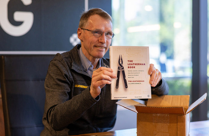 Translation missing: en.Tim Leatherman smiles while holding up a copy of The Leatherman Book beside an open shipping box on a conference table