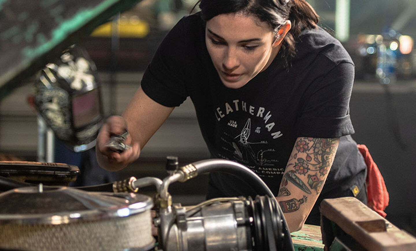 Woman mechanic wears black Leatherman PST Heritage T-Shirt while working under the hood of a vehicle.