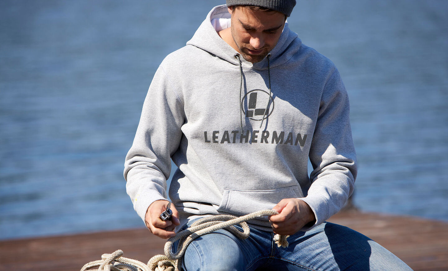 Mean wearing dark gray beanie, gray Leatherman logo hoodie on deck cutting rope with Leatherman tool 