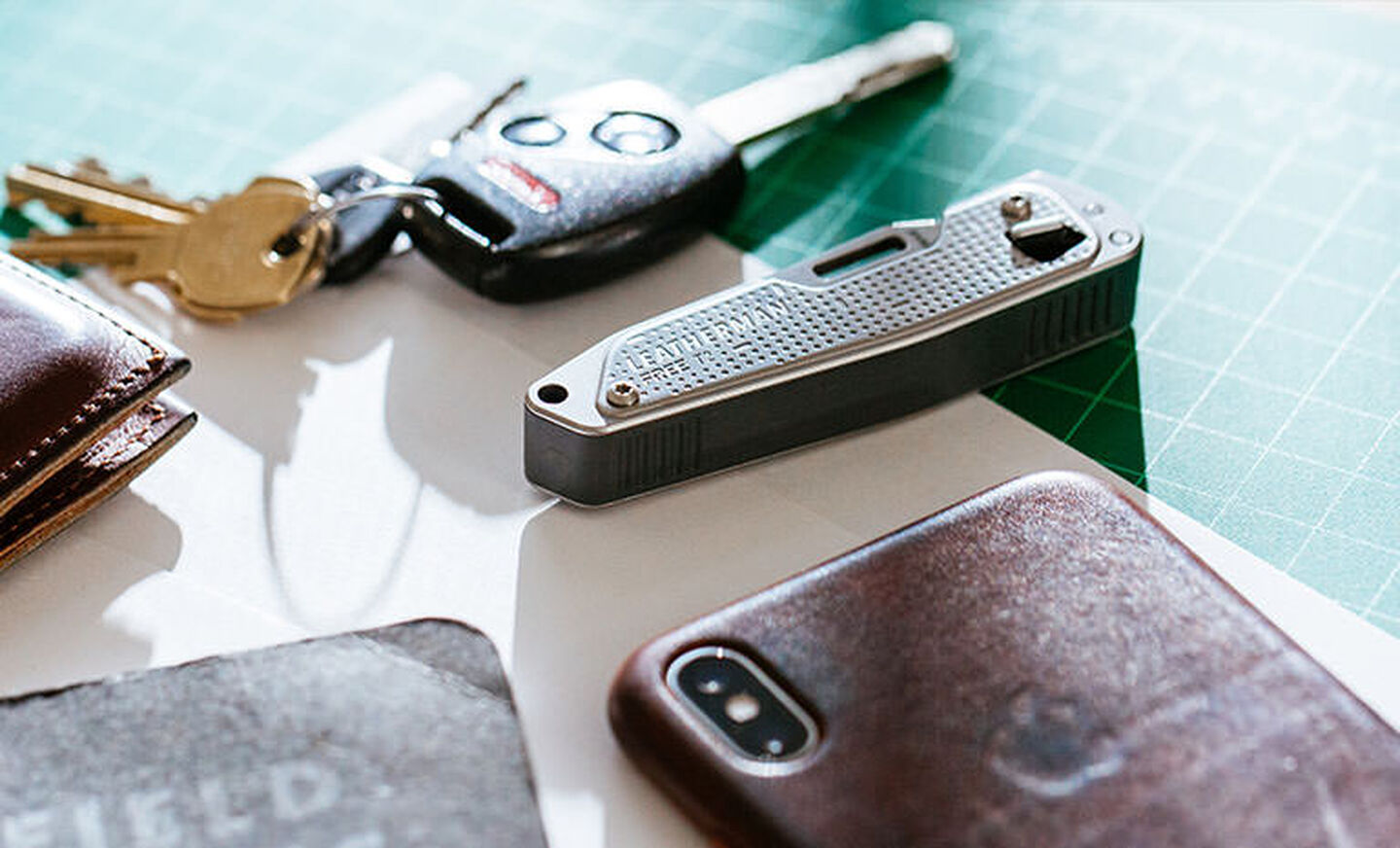 Brown Leatherman Wallet, car keys, Leatherman T2, gray note pad, and Iphone on top of white piece of paper placed on green grid table