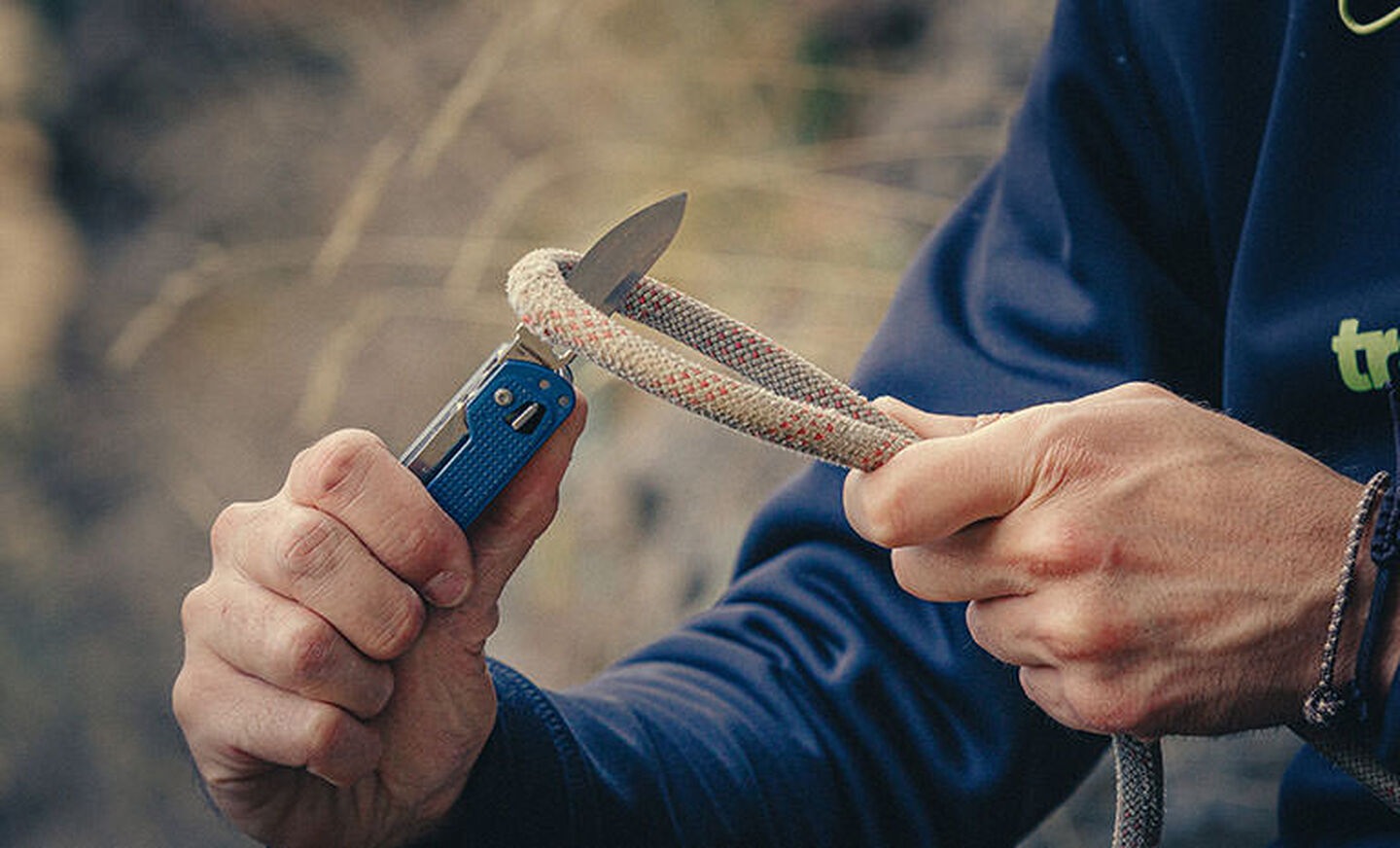 Man in blue long sleeve T shirt using knife blade on Leatherman Free T4 to custom thick beige and orange rope