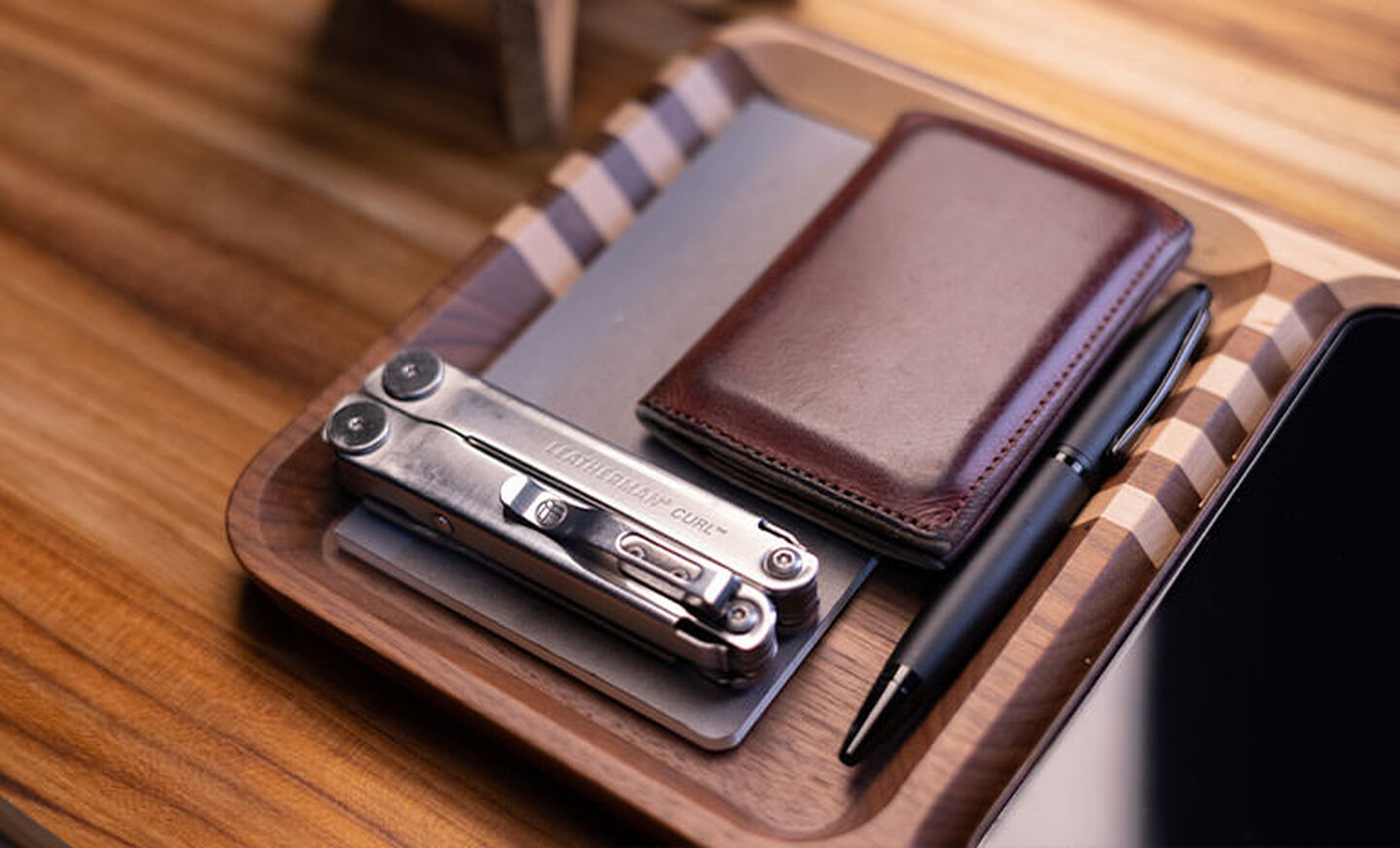 Leatherman Curl, dark brown leather wallet and black pen on top gray notepad nicely arrange on wooden tray