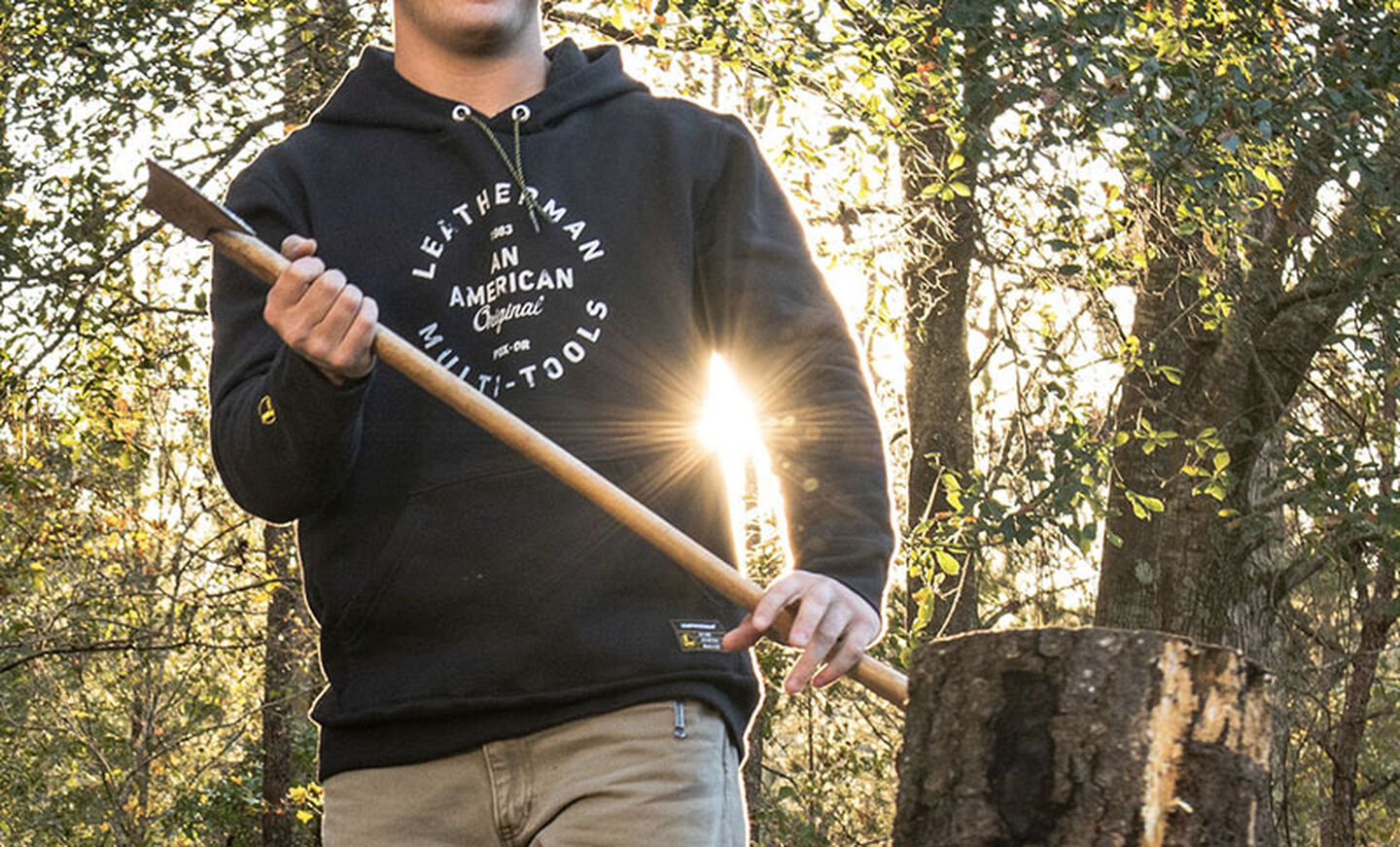 Man wearing black Leatherman Heritage Badge Pullover Hoodie chops firewood with an axe.
