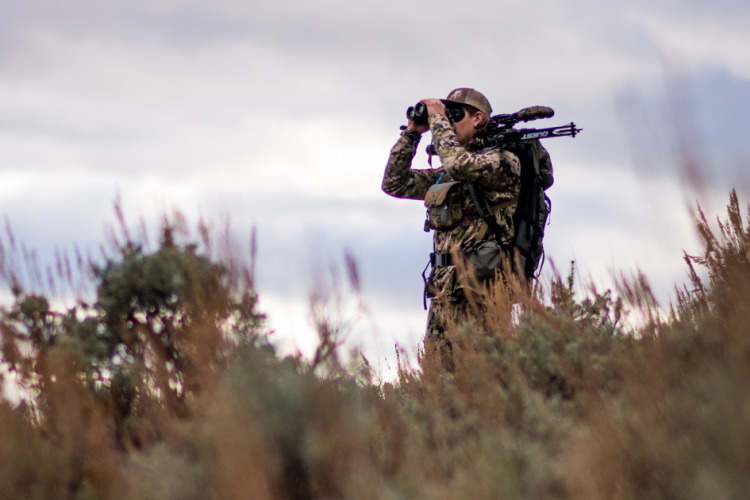 Twee jagers met geweren bovenop een berg met een verrekijker om herten of elanden te zoeken.