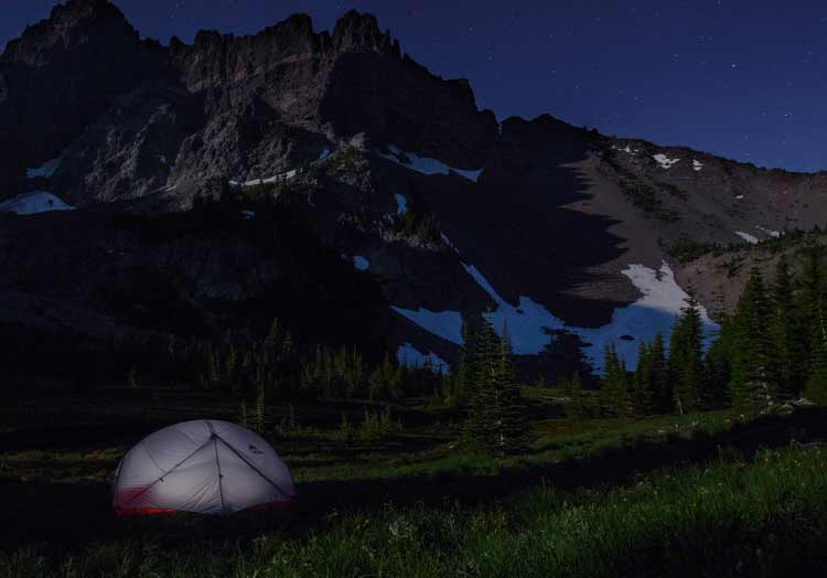 Tente de camping installée sur les hauteurs d'une montagne enneigée la nuit, avec étoiles scintillantes dans le ciel.