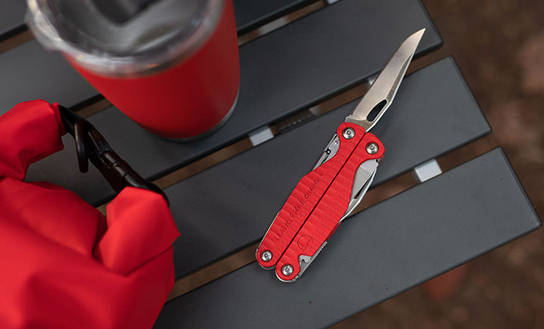 A red Leatherman Charge+ G10 layiing on a table with the knife blade open next to a cup and a pouch